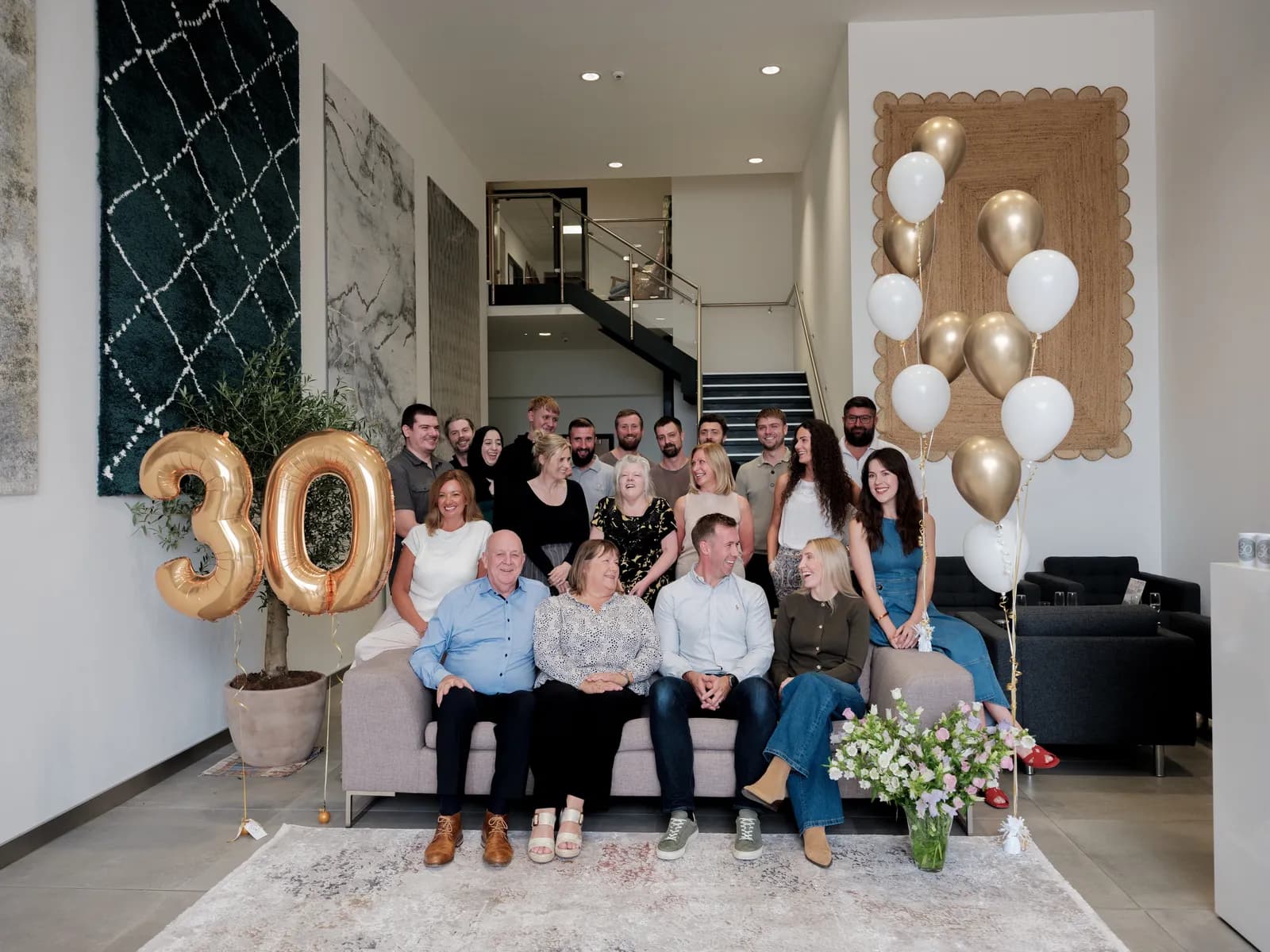 A large group of people of varying ages gather indoors for a 30th-anniversary celebration, with some seated on a sofa and others standing behind them, all surrounded by golden number 30 balloons and additional gold and white balloons.
